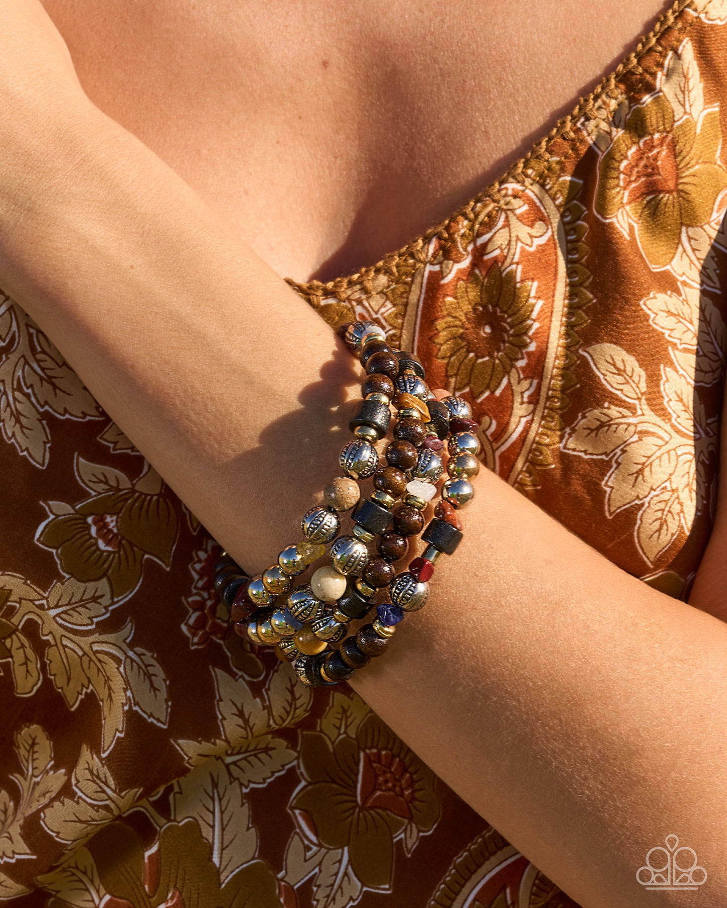 A close-up of a person's wrist wearing a stack of bohemian beaded bracelets, styled with a brown and gold floral-patterned top.