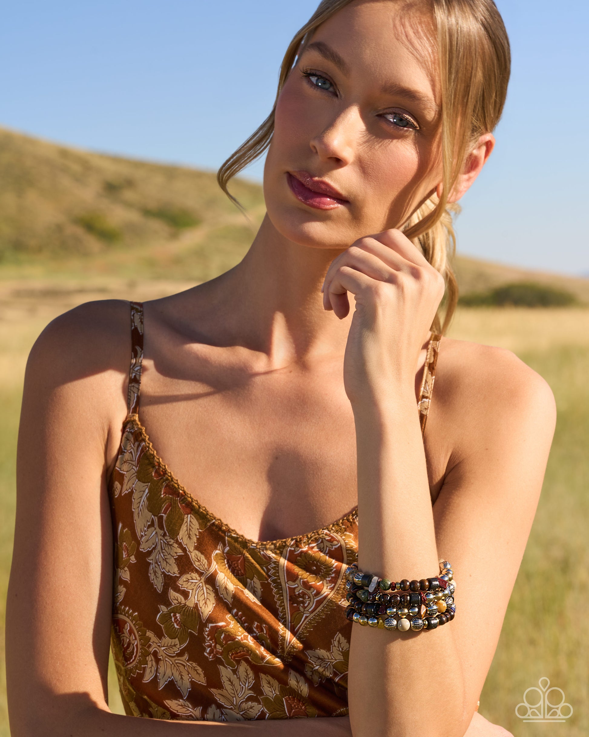 A woman outdoors in a grassy field wearing a brown and beige floral print camisole top and stacked beaded bracelets.