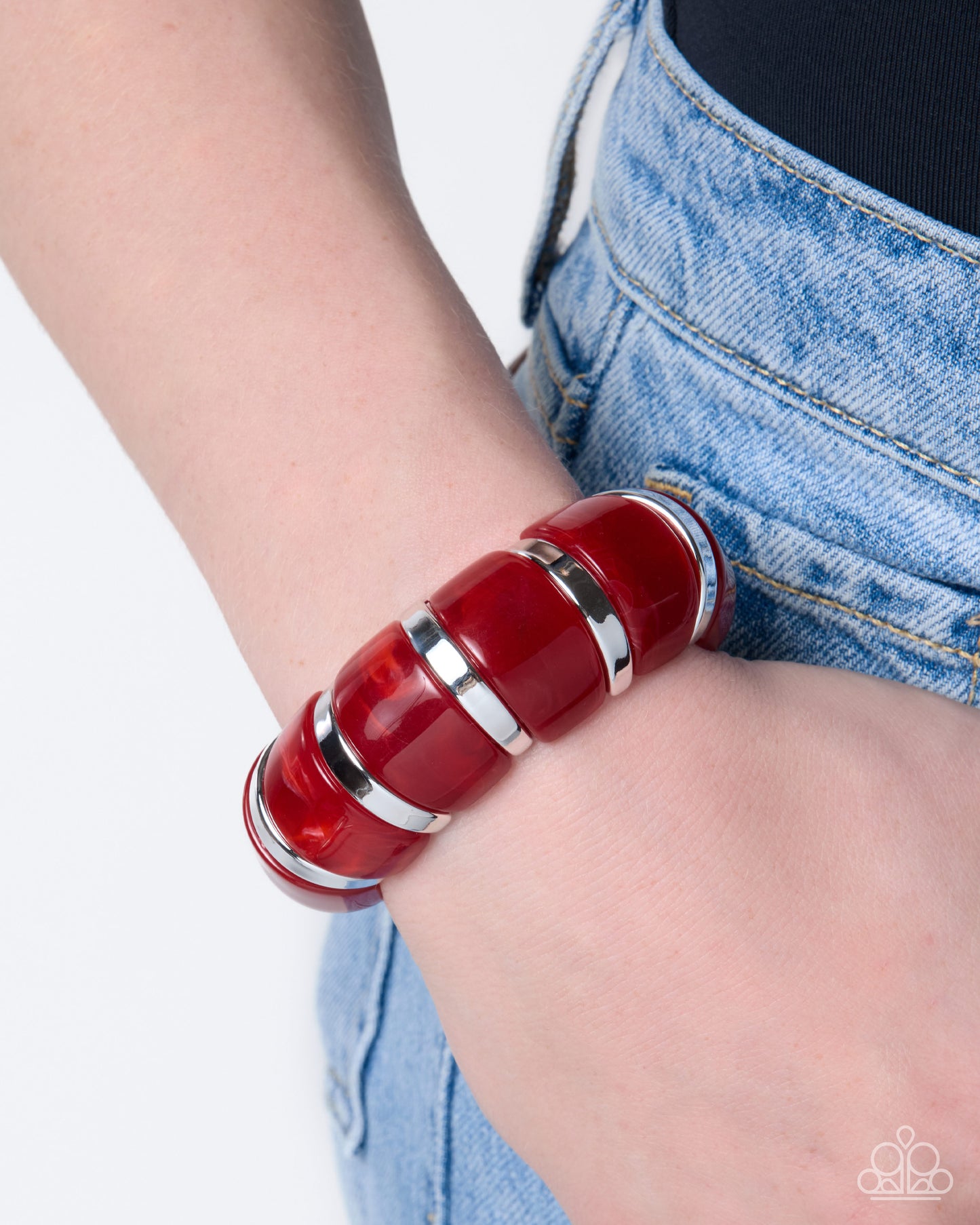 A close-up of a person's wrist wearing a chunky, segmented red resin bracelet with polished silver metal bands, paired with light wash denim jeans.