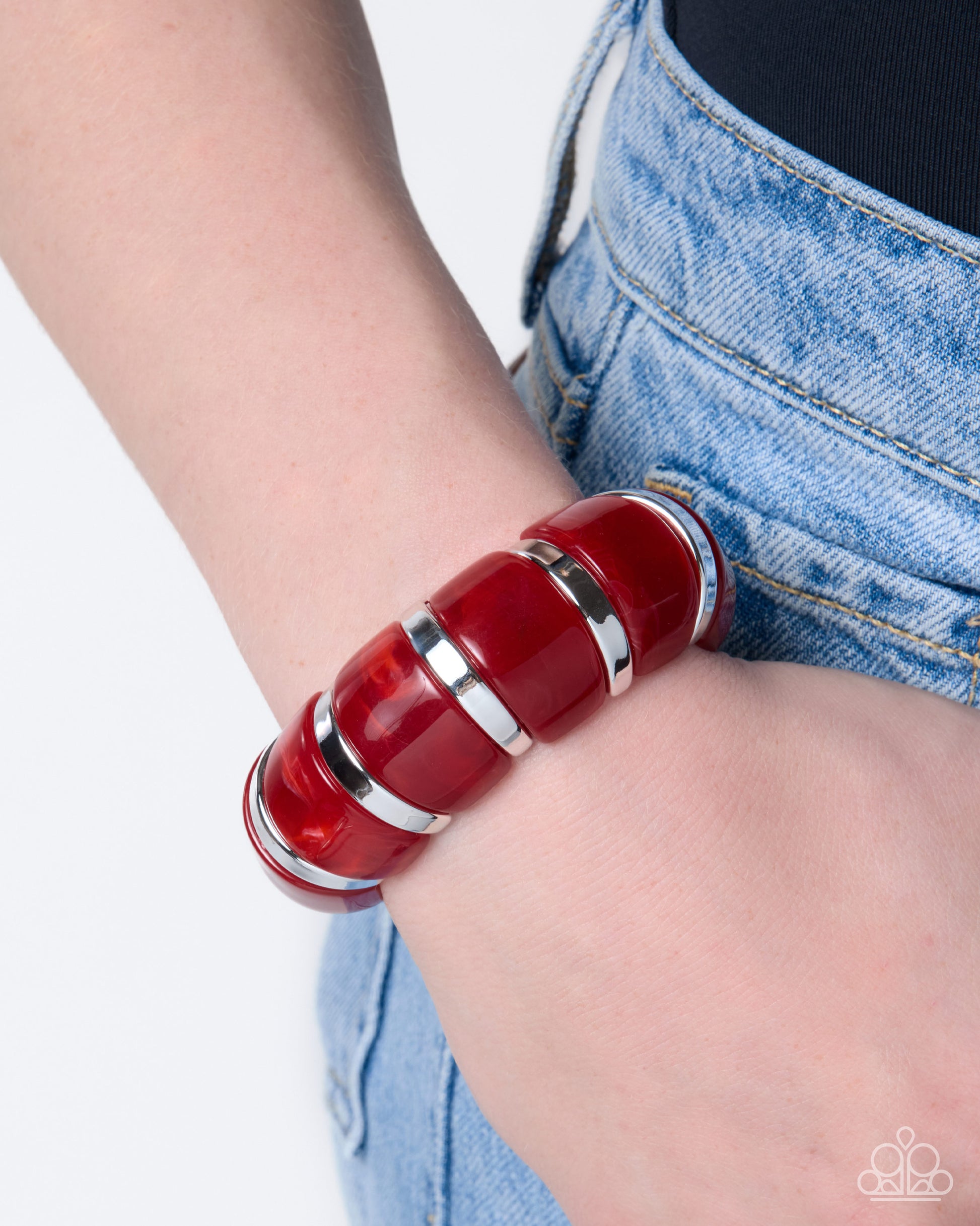 A close-up of a person's wrist wearing a chunky, segmented red resin bracelet with polished silver metal bands, paired with light wash denim jeans.