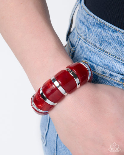 A close-up of a person's wrist wearing a chunky, segmented red resin bracelet with polished silver metal bands, paired with light wash denim jeans.
