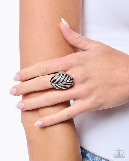 A close-up of a person's hand resting on their forearm, wearing a large oval ring with a black and white zebra-stripe pattern. The background is a clean white studio setting.