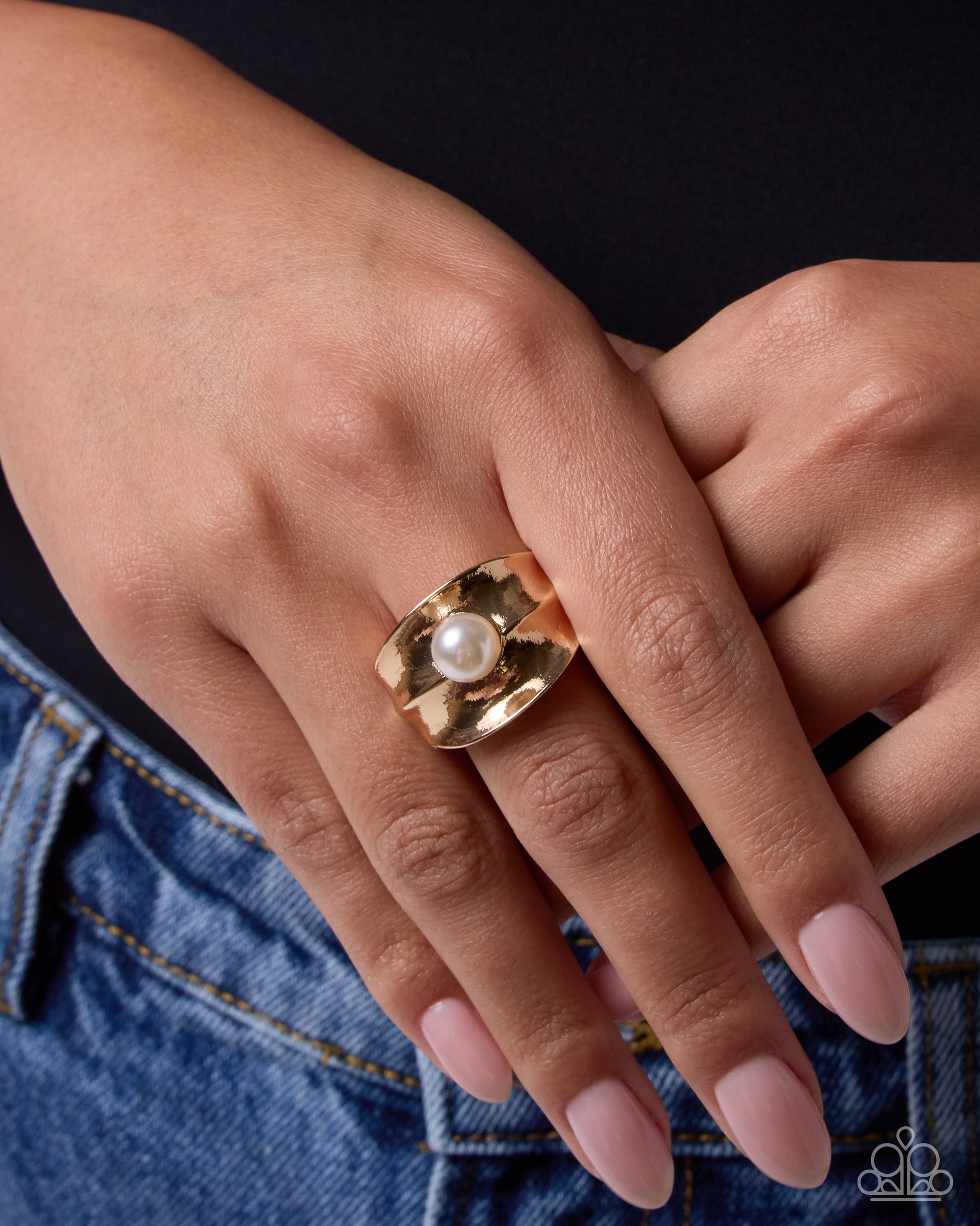 A close-up of a person's hand wearing a wide gold ring with a central white pearl, paired with light pink manicured nails and blue denim jeans against a dark background.