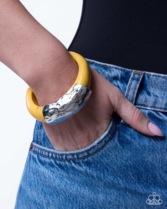 A close-up of a person wearing a chunky bangle bracelet with a yellow top half and a textured silver bottom half, paired with blue jeans.
