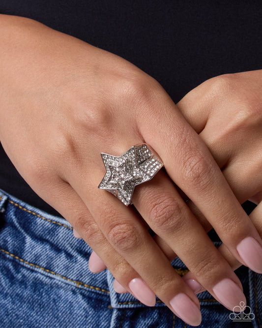 A close-up lifestyle shot of a person's hand wearing a large, star-shaped silver ring paved with clear crystals, styled with a black top and blue denim jeans.