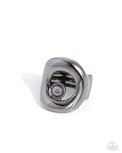 A dark gray metallic ring with a unique sculptural, organic shape and a textured circular detail, shown on a clean white background.