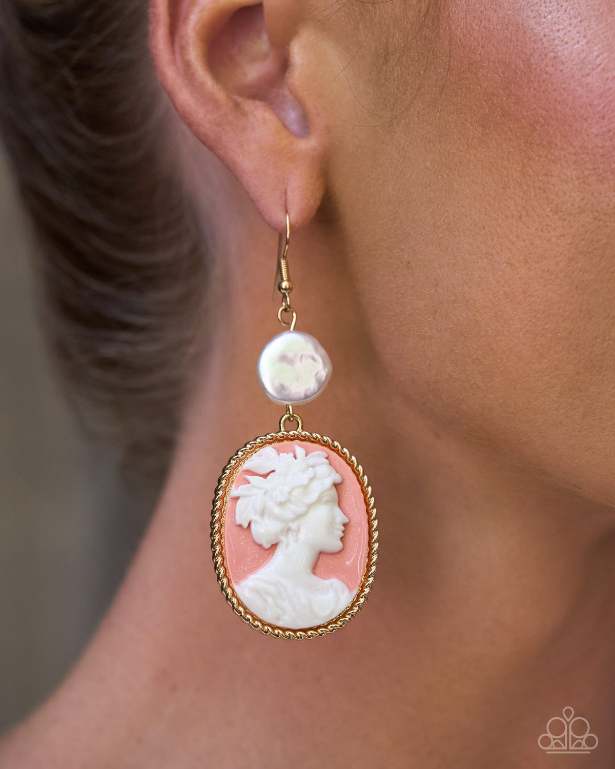 A close-up of a model's ear wearing a gold dangle earring with a round white pearl and an oval carved cameo-style face in white on a pink background.