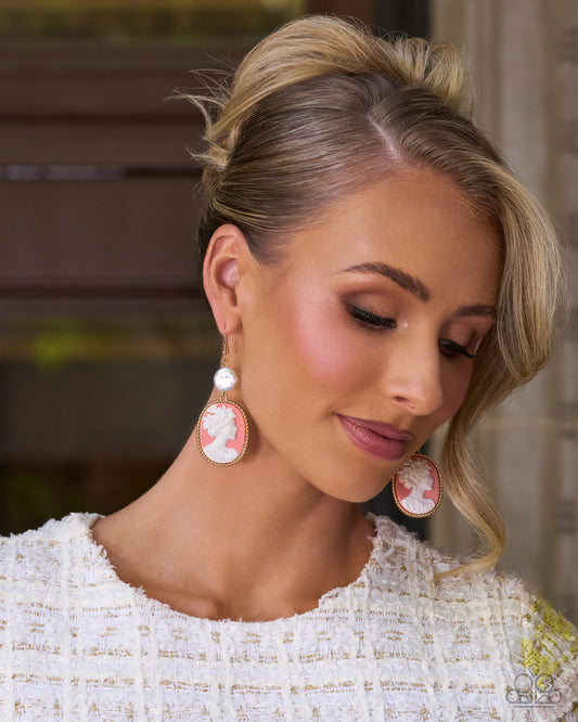 A close-up lifestyle shot of a woman with blonde hair styled in an updo, wearing a white textured tweed top and matching white and pink cameo-style drop earrings.
