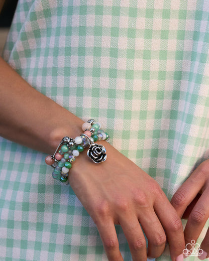 A close-up of a person's wrist wearing a stack of beaded bracelets, including a black and white rose charm, on a mint green and white gingham dress.
