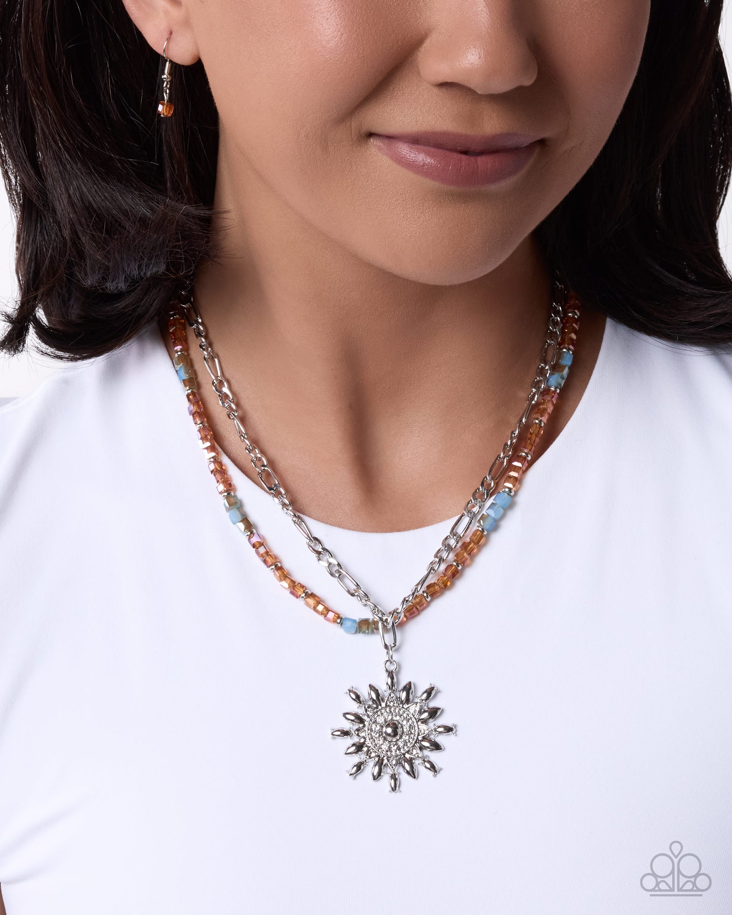 A close-up of a model wearing a multi-strand necklace featuring silver chains, orange and light blue beads, and a silver sunburst pendant, paired with a white t-shirt.