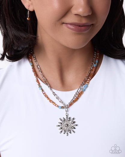 A close-up of a model wearing a multi-strand necklace featuring silver chains, orange and light blue beads, and a silver sunburst pendant, paired with a white t-shirt.