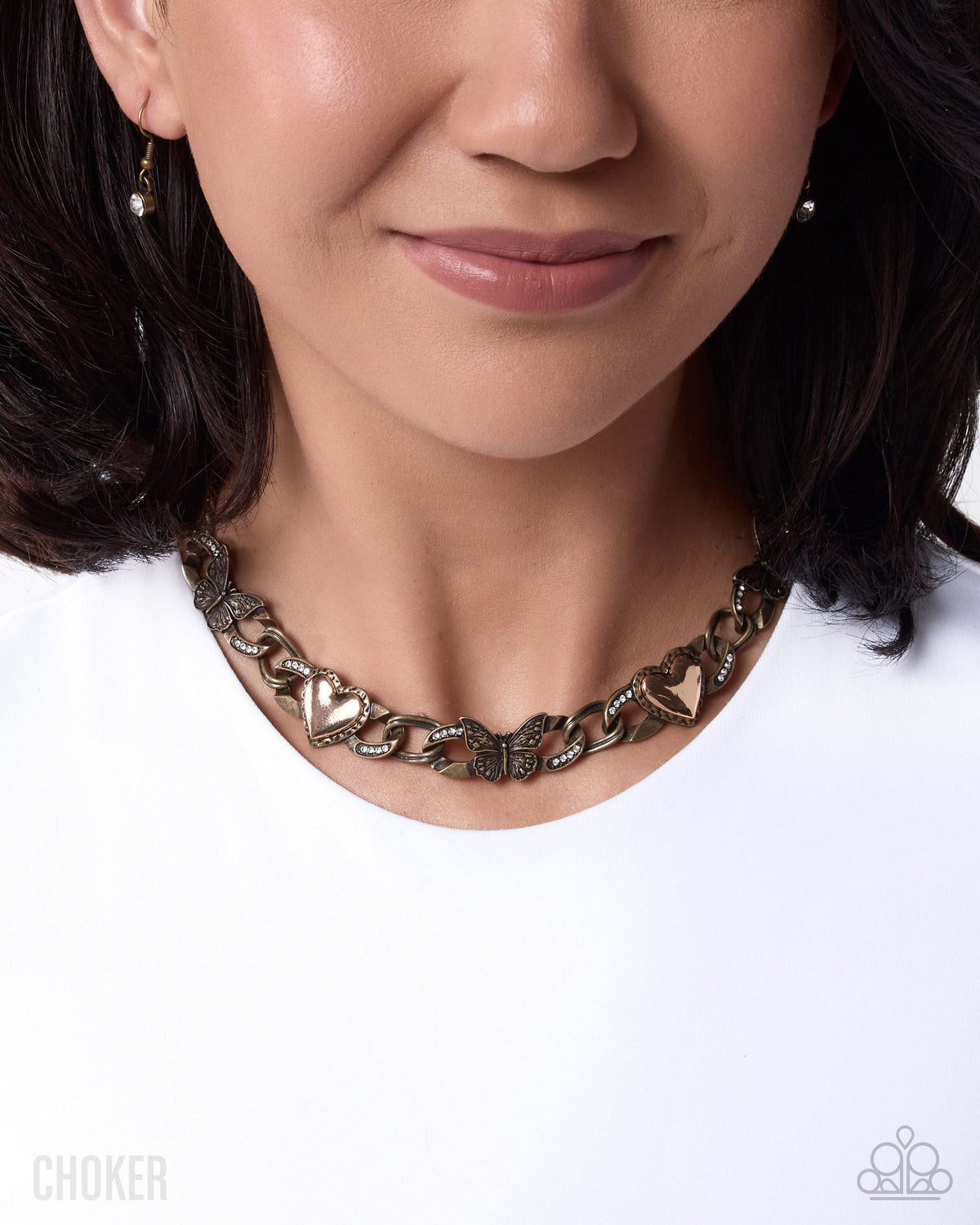 A close-up of a woman wearing a Choker brand necklace with interlocking heart links in an antique bronze finish, paired with small dangling earrings and a white top.