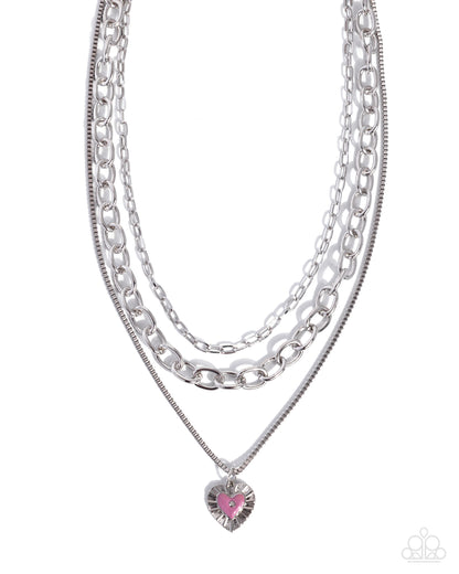 A layered silver-tone necklace featuring three distinct chain styles and a dangling heart-shaped charm with a pink center, shown against a plain white background.