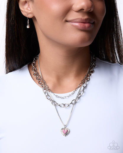 A close-up of a woman wearing a layered silver necklace set with a heart-shaped pendant featuring a pink ribbon in the center, styled with a white crewneck top.