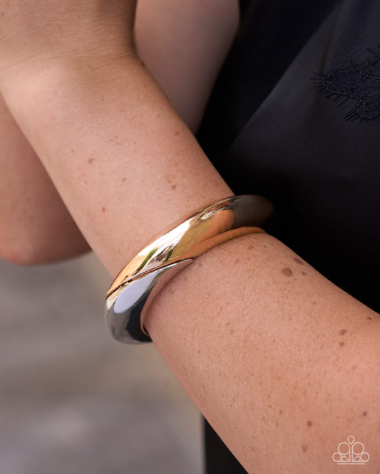 A close-up of a person's wrist wearing a polished two-tone metal bangle bracelet, showing one gold-toned band and one silver-toned band, with a black embroidered top in the background.