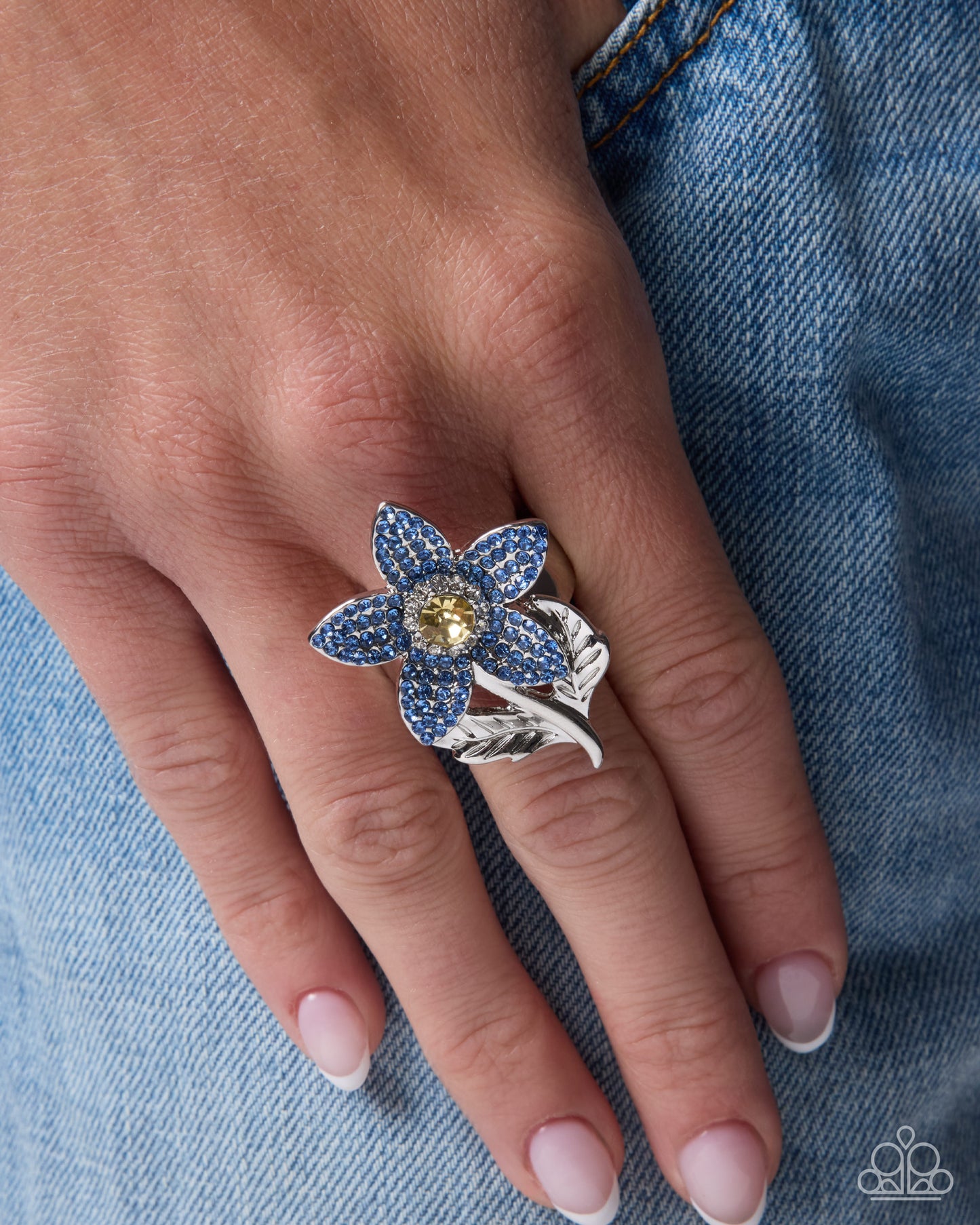 A close-up of a hand wearing a floral ring with blue pavé petals, a yellow center stone, and a silver leaf-shaped band, set against blue denim.