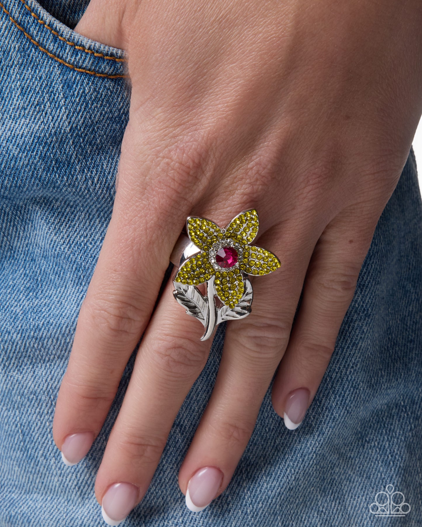 A close-up of a hand wearing a silver ring designed as a flower with yellow pavé petals and a red center stone, resting against blue denim.