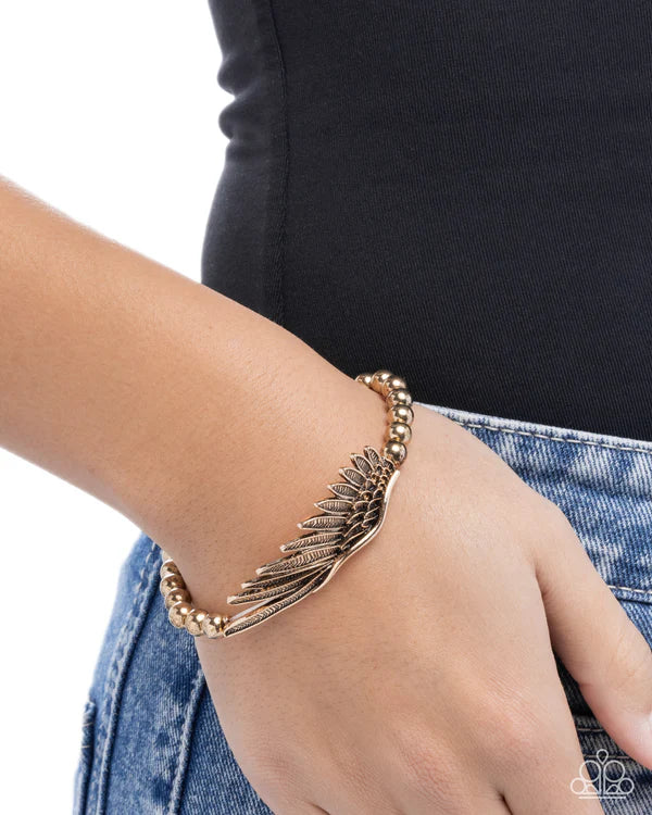 Gold wing-shaped bracelet on a wrist with a black sleeve and blue jeans background