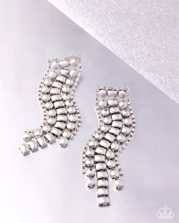 A pair of silver-toned statement earrings with cascading rows of clear rhinestones and teardrop charms, displayed on a light gray surface with a textured silver diagonal background.