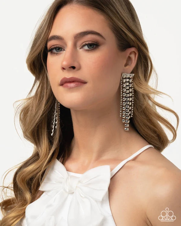 A close-up of a woman wearing long, dangling silver-toned statement earrings with multiple rows of clear rhinestones, paired with a white bow-neck top against a clean white background.