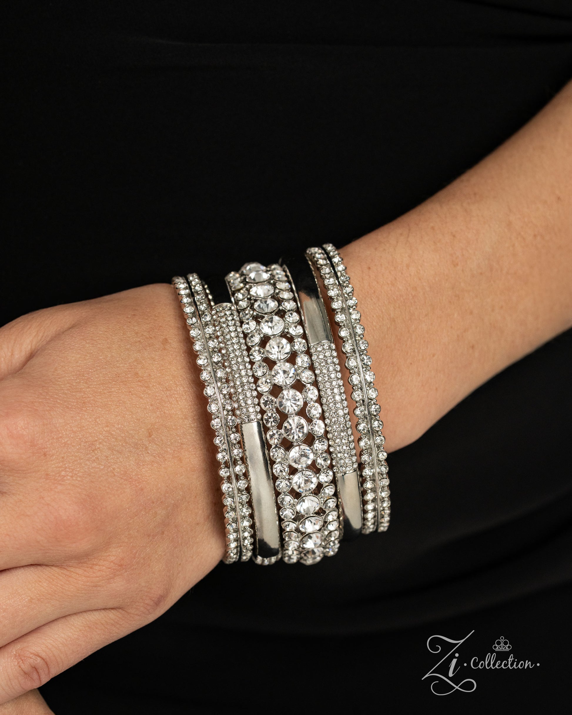 A close-up of a person's wrist wearing a stack of wide silver bangles adorned with clear rhinestones and crystal-like stones, set against a black top background. A 'Zi Collection' logo watermark is visible in the bottom right corner.