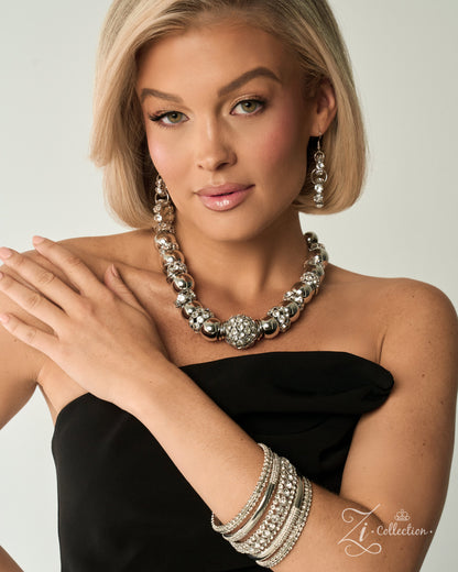 A blonde model wearing a chunky silver statement necklace, matching drop earrings, and stacked silver bangles, paired with a black strapless top against a light gray background.