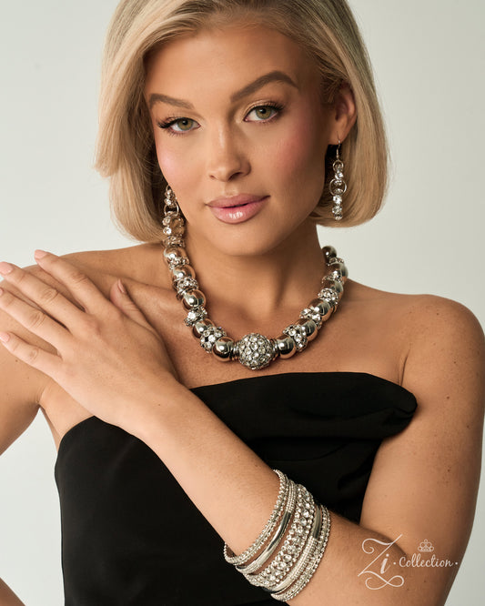 A blonde model wearing a chunky silver statement necklace, matching drop earrings, and stacked silver bangles, paired with a black strapless top against a light gray background.