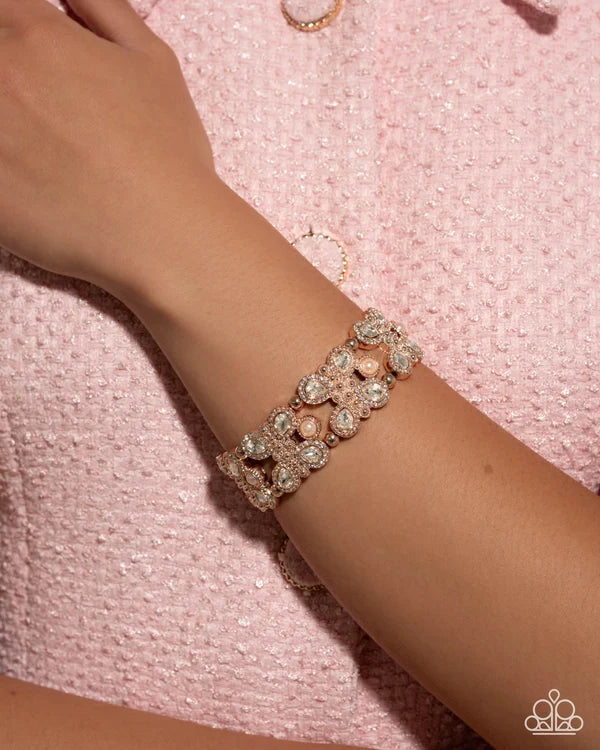 A close-up of a model's wrist wearing a wide, ornate bracelet with clear crystals and pale pink pearls on a textured light pink fabric background.
