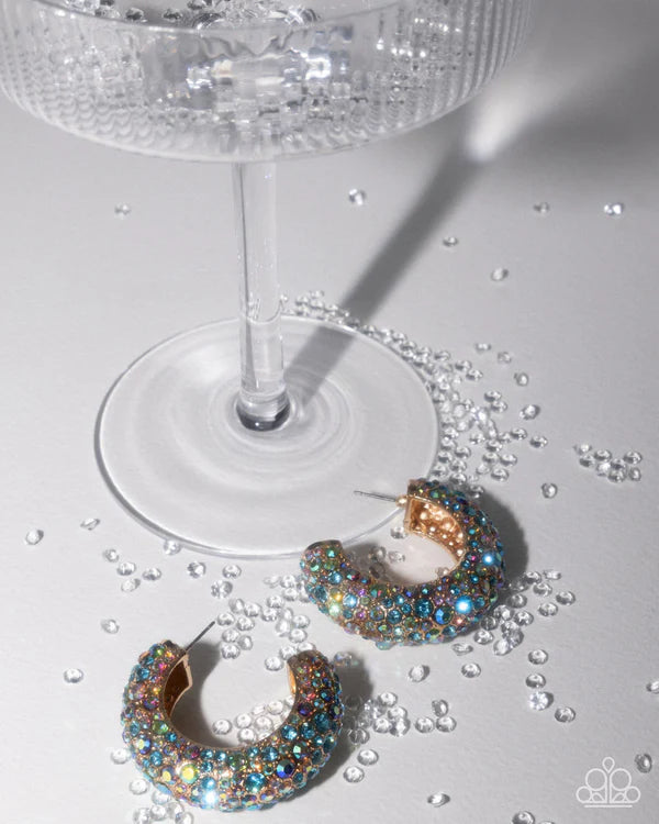 Multicolored hoop earrings on a white surface with a glass and scattered rhinestones.