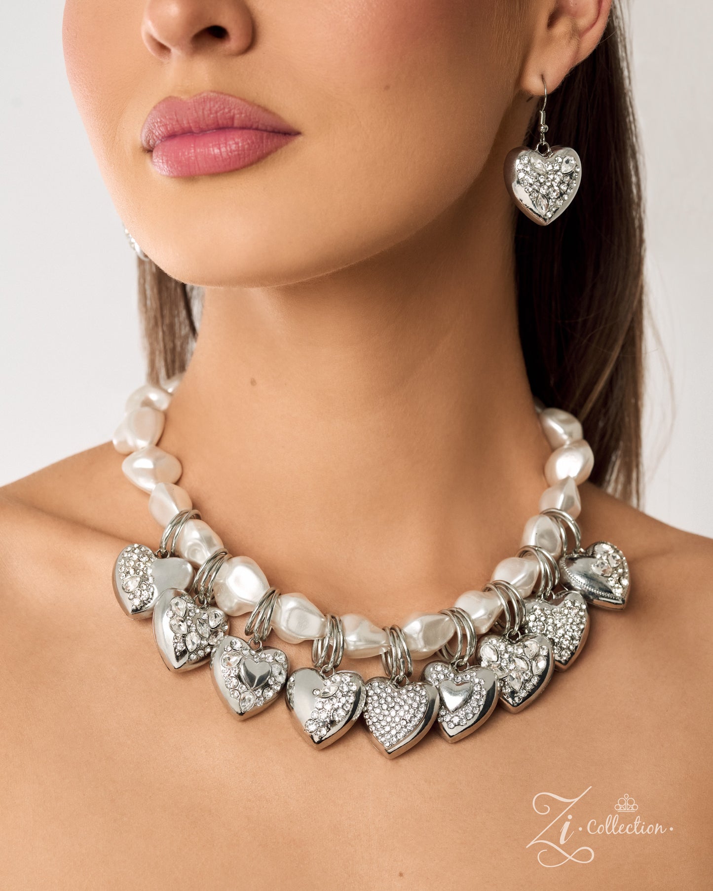 A close-up of a woman wearing a silver heart-studded statement necklace and matching heart drop earrings. The necklace features silver heart charms, some encrusted with clear rhinestones, alongside white faux pearls.