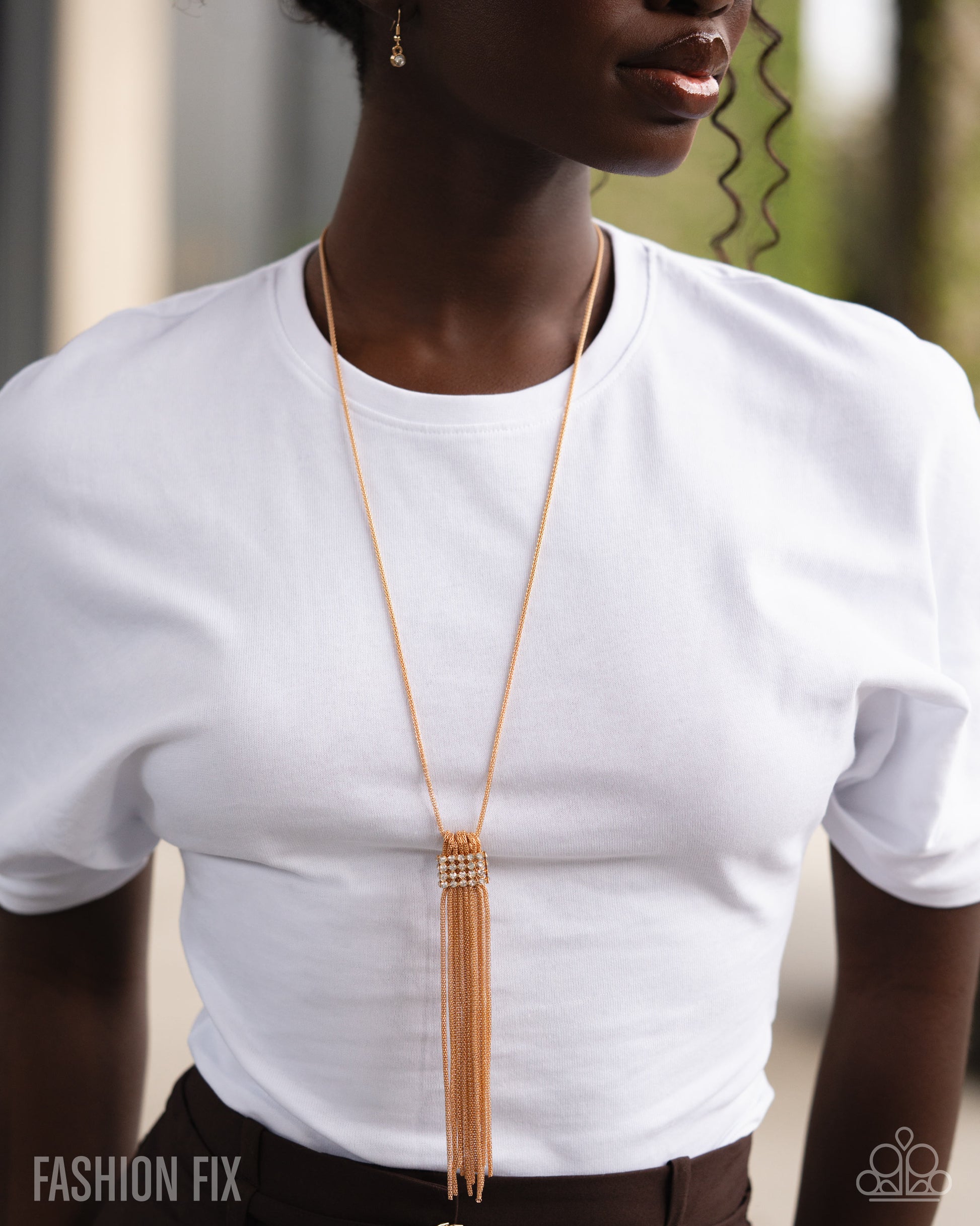 Person wearing a long gold necklace with tassels on a white shirt.