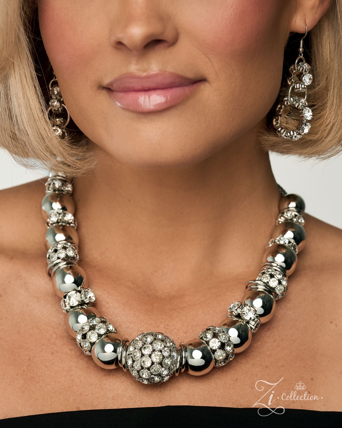 A close-up of a model wearing a chunky silver statement necklace with large polished beads, crystal-encrusted details, and a central round crystal cluster, paired with matching dangling earrings.