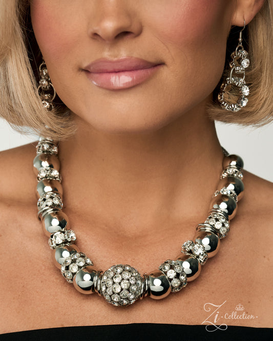 A close-up of a model wearing a chunky silver statement necklace with large polished beads, crystal-encrusted details, and a central round crystal cluster, paired with matching dangling earrings.