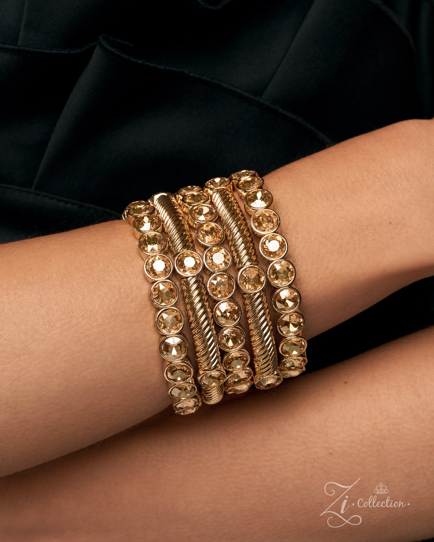 A stack of five gold-toned bangle bracelets worn on a person's wrist, featuring round clear rhinestones and twisted rope details, set against a dark black fabric background.