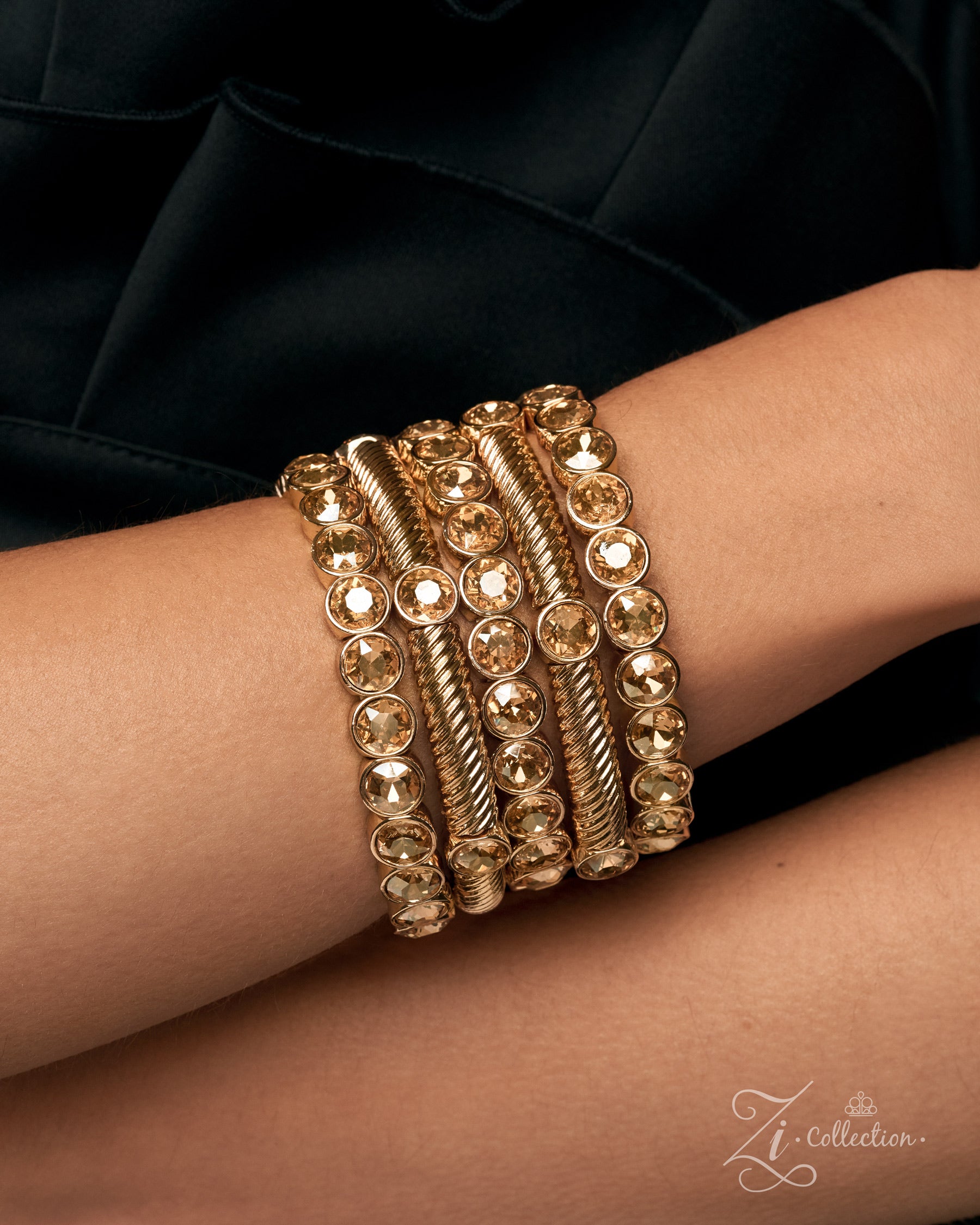 A stack of five gold-toned bangle bracelets worn on a person's wrist, featuring round clear rhinestones and twisted rope details, set against a dark black fabric background.