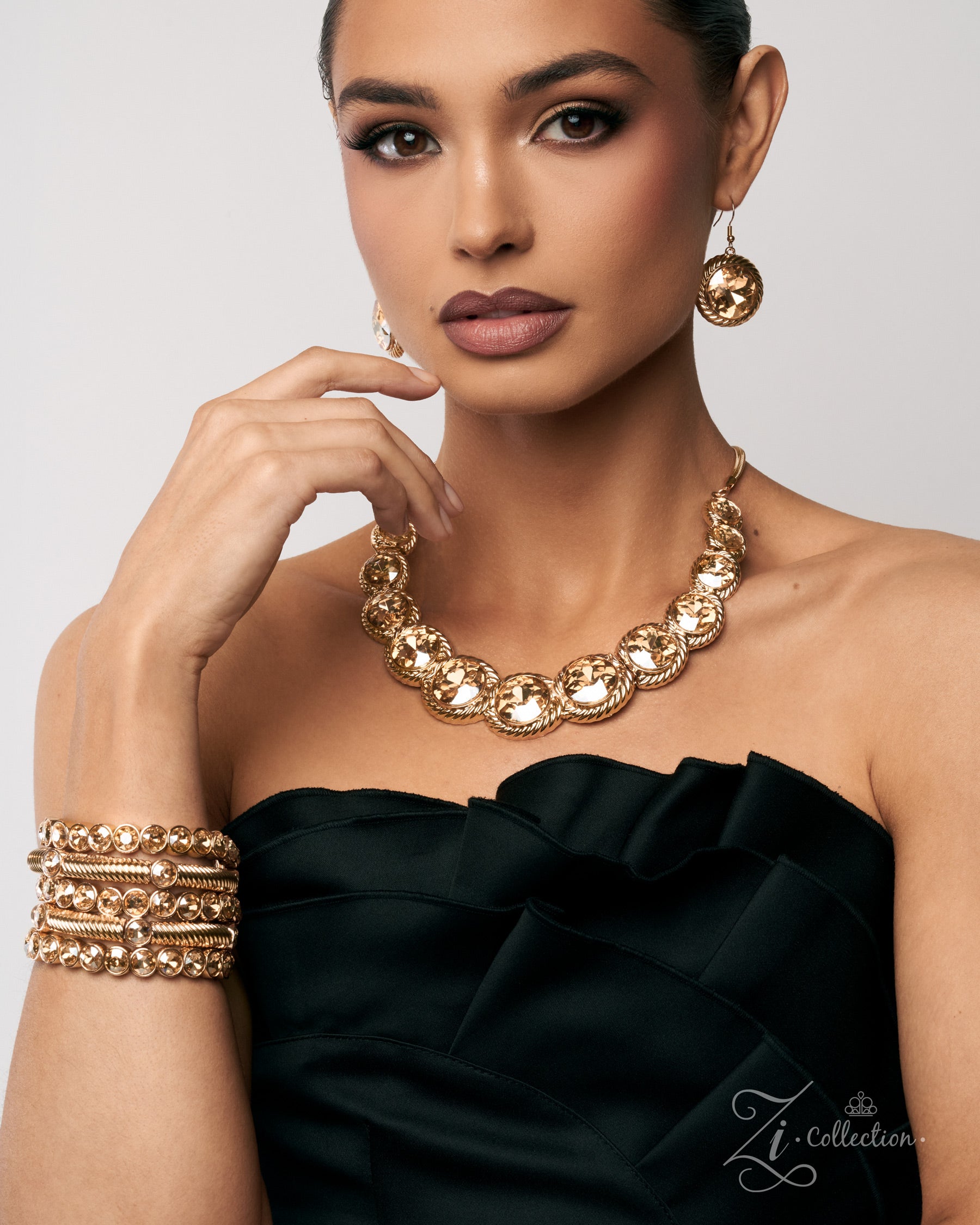 A woman models a gold-tone jewelry set from Zi Collection, featuring a chunky link collar necklace, matching drop earrings, and stacked bangles, paired with a black strapless ruffled top against a light gray background.