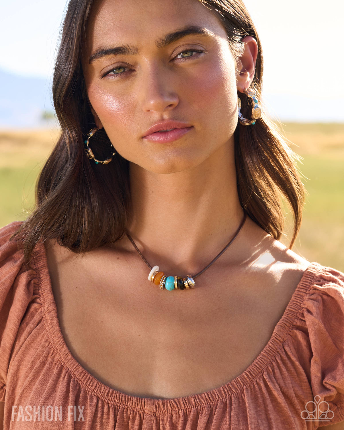 A woman outdoors wearing a beaded necklace with turquoise, white, and black beads on a dark cord, paired with large gold hoop earrings and a dusty rose top.