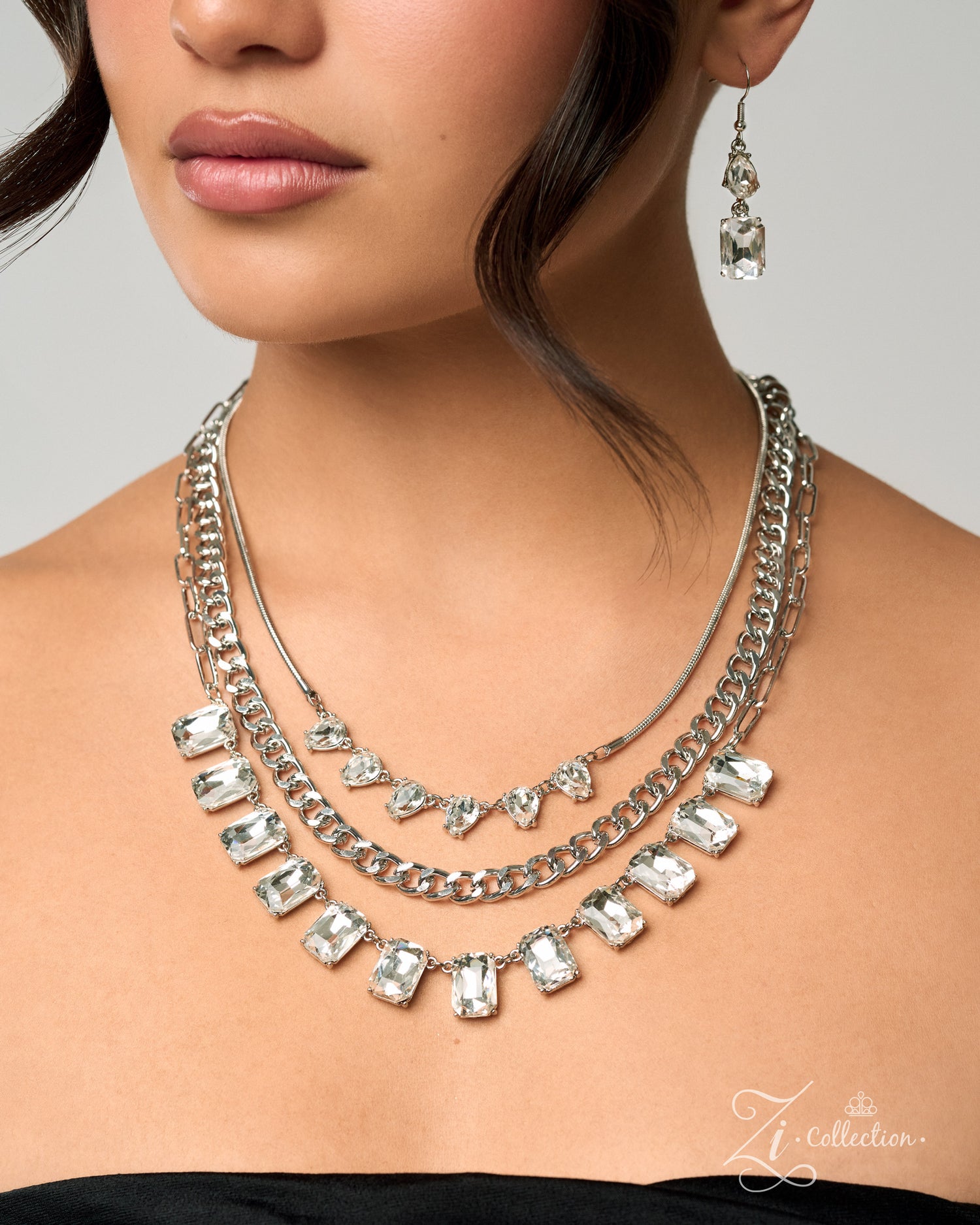 A close-up of a model wearing a multi-layered silver necklace with clear rectangular and heart-shaped rhinestones, along with matching dangling earrings, against a light gray background.