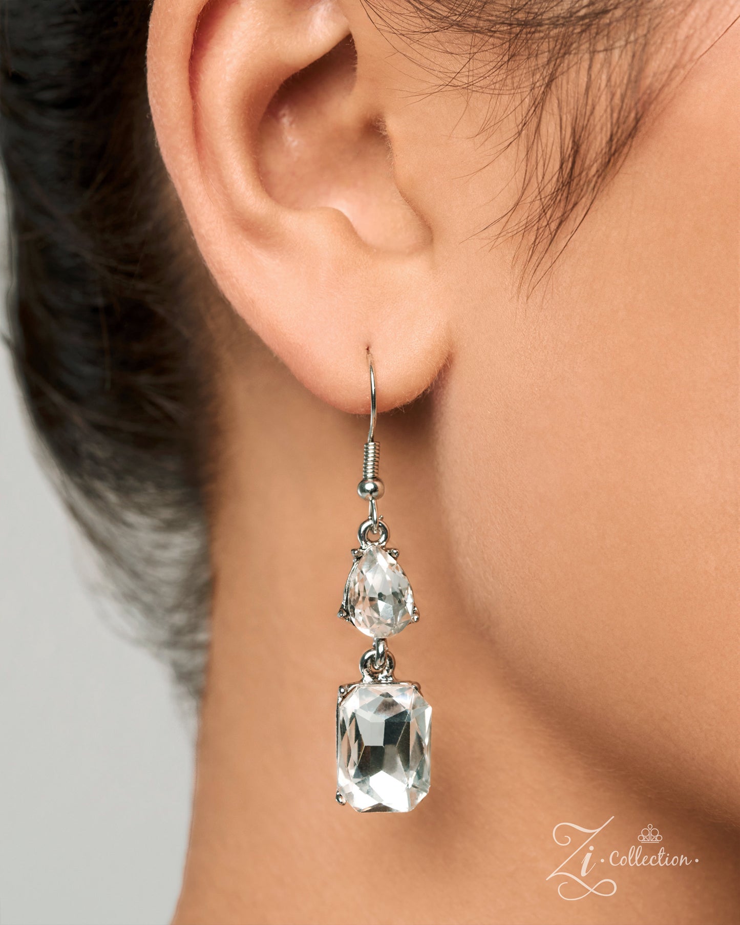 A close-up of a person's ear wearing a silver-tone drop earring with two clear faceted gemstones, shown against skin and dark hair. A 'Zi Collection' watermark is visible in the lower right corner.