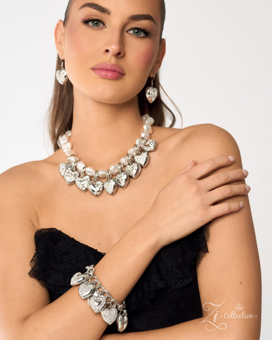 A woman models a silver-toned jewelry set featuring a heart-shaped pendant necklace, matching drop earrings, and a heart-link bracelet, styled with a black strapless top against a light gray background.